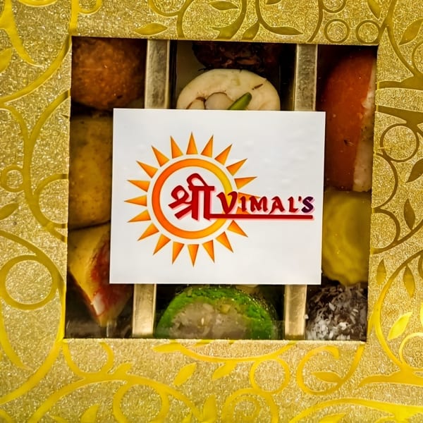 A gold box of assorted Indian sweets with a "Shree Vimal's" logo, next to a gold fork.