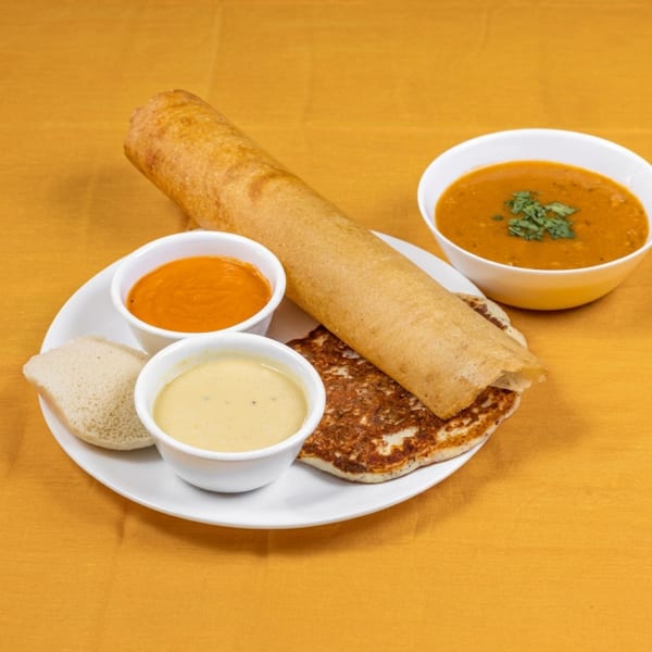 Dosa, uttapam, idli, sambar, and two chutneys on a white plate against a yellow background.
